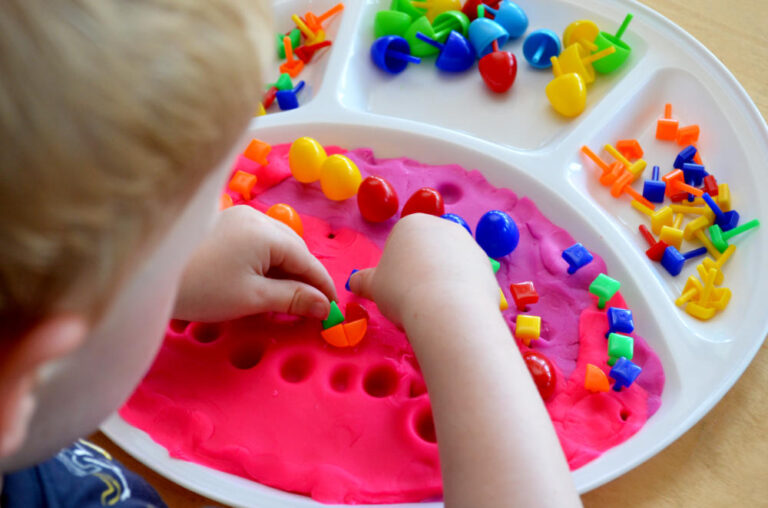 child sticking peg board pegs into tray of playdough