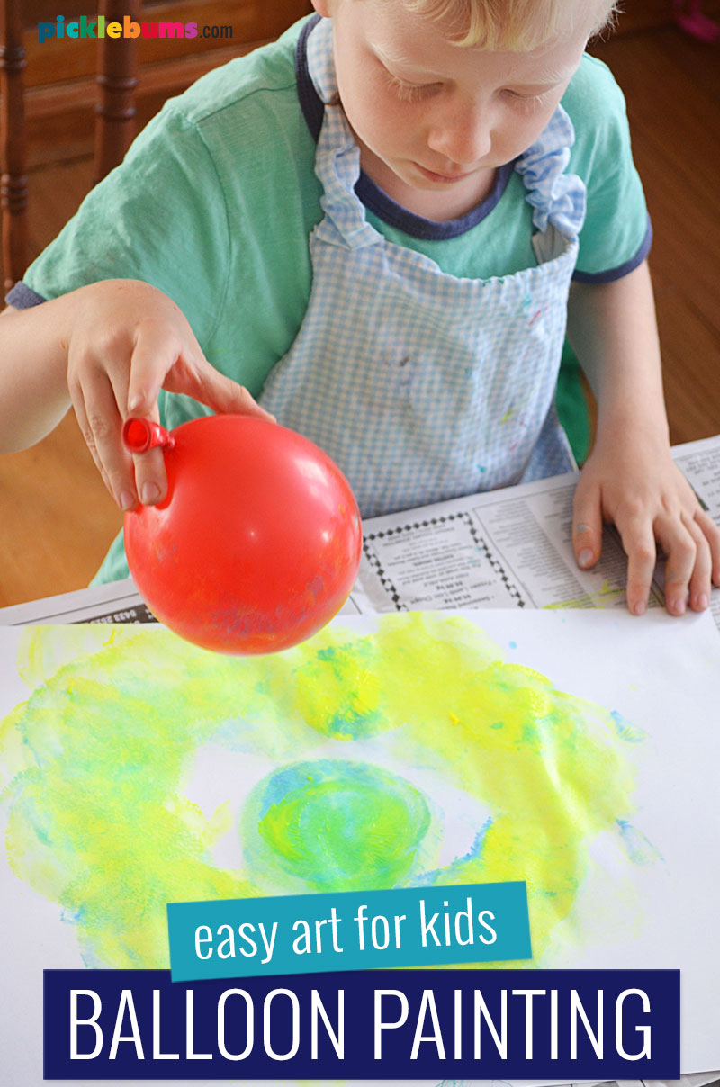child wearing an apron holding a red balloon and painting with it by printing it onto paper