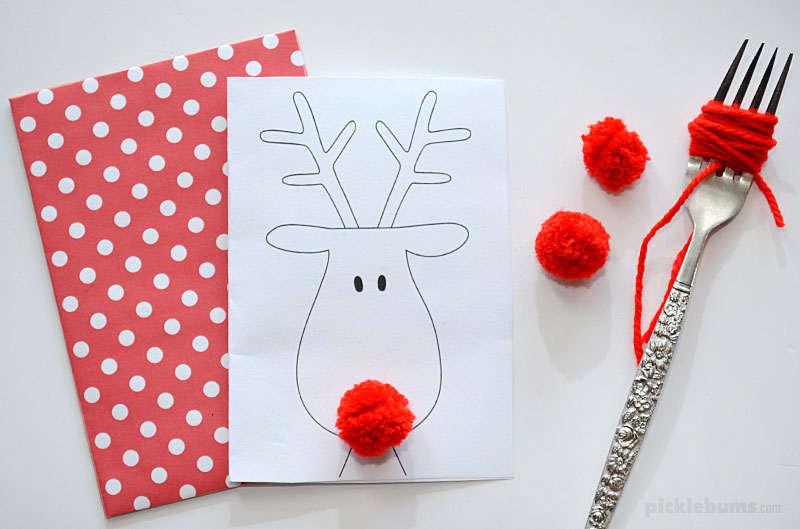 reindeer card with matching red spotty envelope next to fork and red wool pom poms