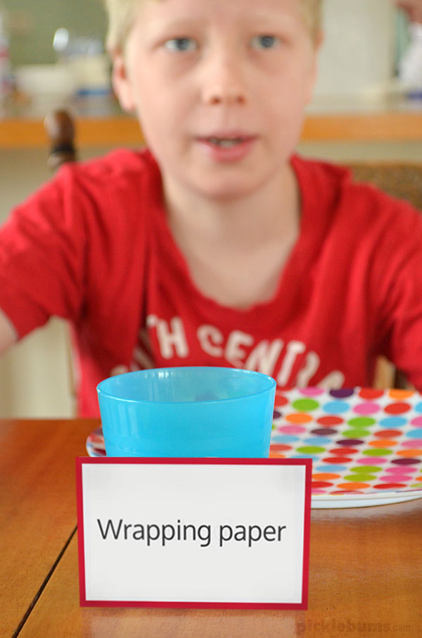 Boy sitting at the dinner table with card saying 'wrapping paper' leaning on his cup